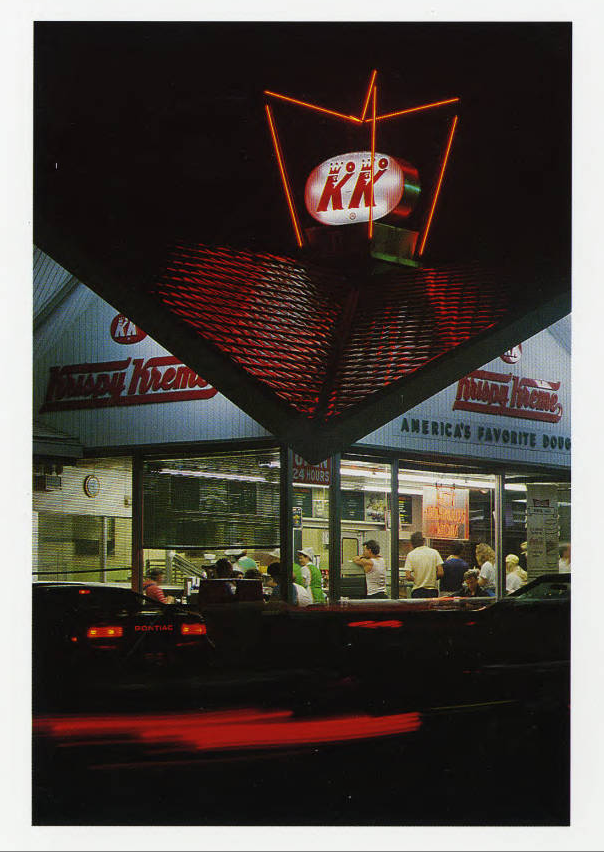 Exterior of Krispy Kreme store at night, with neon sign lit and the warm interior full of people beckoning customers inside.