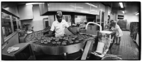 Black and white photo of an African American man wearing a Krispy Kreme hat stands behind a conveyer belt with Krispy Kreme doughnuts.
